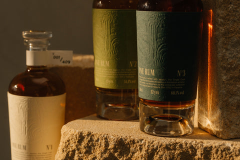 Drei ONE RUM Flaschen mit stilvoll gestalteten Etiketten „No. 1“, „No. 2“ und „No. 3“ auf einem sandfarbenen Stein arrangiert. Diese limitierte Single Cask Auswahl betont die Exklusivität und Herkunft dieser Rums.