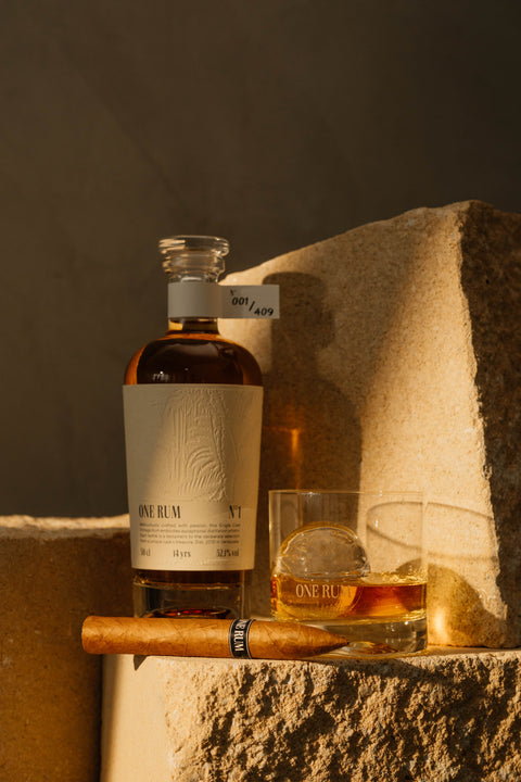ONE RUM No. 1 Flasche neben einem Tumblerglas mit Eis und einer ONE RUM Zigarrenbeilage, stilvoll auf Stein inszeniert. Die Szene spiegelt den Luxus und die Genusswelt von hochwertigem Single Cask Rum wider.