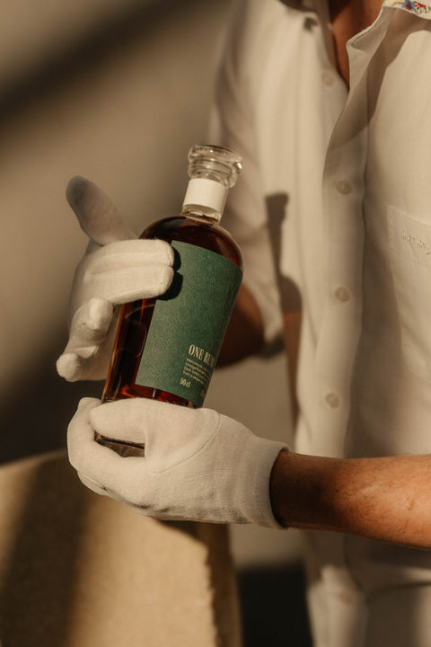 Nahaufnahme einer ONE RUM No. 3 Flasche, die von einer behandschuhten Hand gehalten wird. Die Flasche mit grünem Etikett und tropischem Muster steht für die Exklusivität der Single Cask Kollektion.