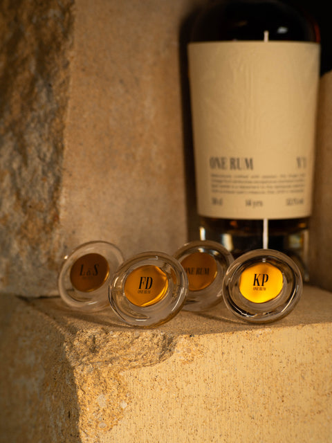Detailaufnahme von personalisierten Glasverschlüssen für ONE RUM Flaschen mit den Initialen "FD", "KP" und "L&S". Die Verschlüsse liegen vor einer ONE RUM No. 1 Flasche und heben die individuelle Gestaltung und Exklusivität dieses Single Cask Rums hervor.