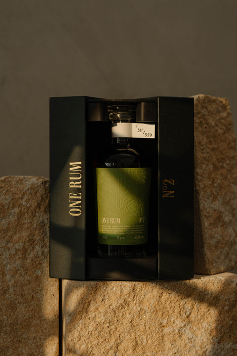 ONE RUM No. 2 in einer edlen schwarzen Geschenkbox mit goldenem Schriftzug, auf einem Steinblock ausgestellt. Diese 13 Jahre gereifte Abfüllung mit einem Alkoholgehalt von 55,3% vol. wird in einem hochwertigen Etui präsentiert.