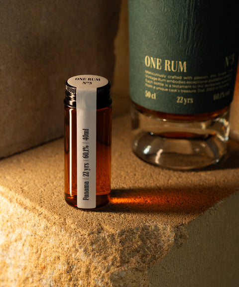 Probefläschchen ONE RUM No3 mit Panama Rum, 22 Jahre gereift und 60,1% Alkoholgehalt, vor einer größeren ONE RUM Flasche präsentiert. Die Szene zeigt den limitierten und exklusiven Charakter dieses Single Cask Vintage Rums.